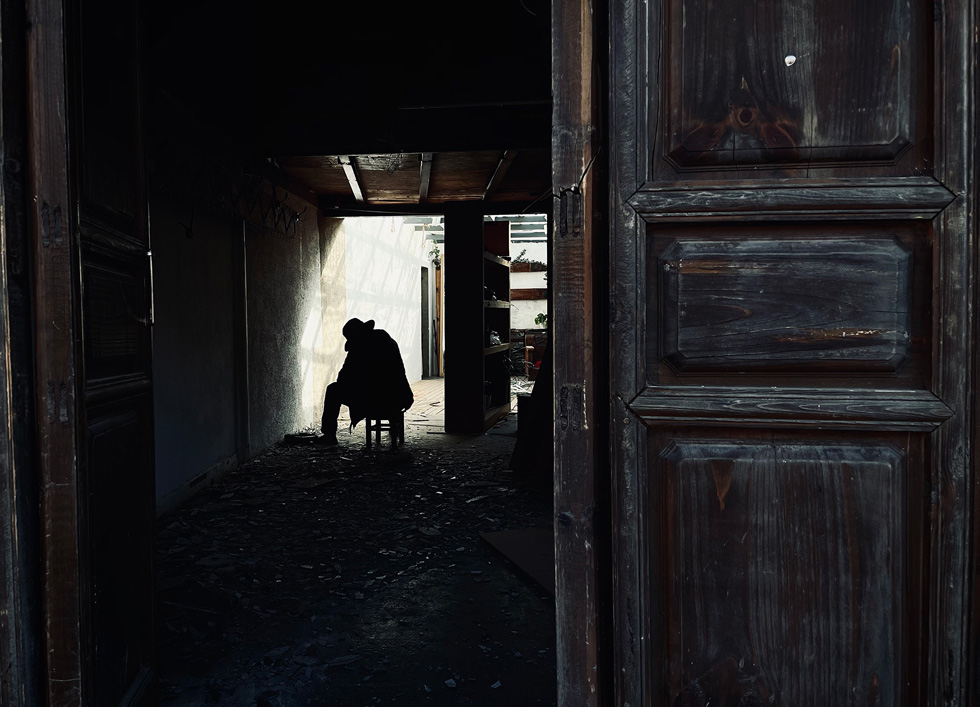 A través de una puerta abierta, se ve una figura sentada entre escombros y sombras.